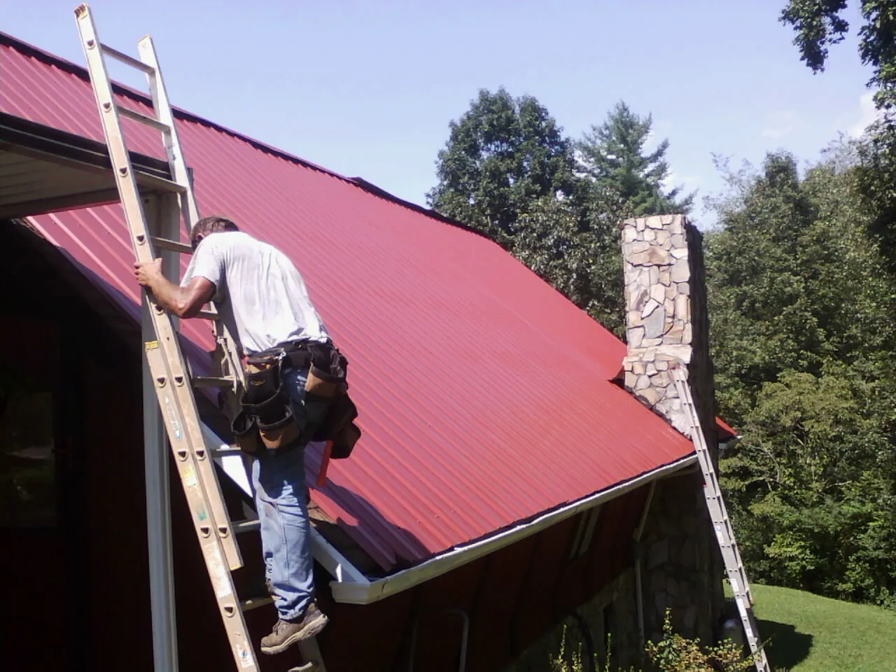 Roofer on a ladder beside a red metal roof and stone chimney — Soffit & Fascia Repair by Roofing Brothers in Aspen
